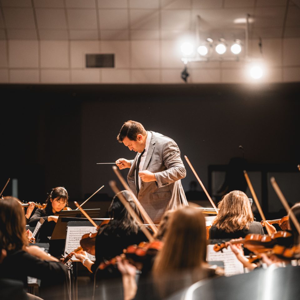 A conductor standing at the center of an orchestra with their head bowed, leading the musicians with raised hands as the ensemble performs around them.